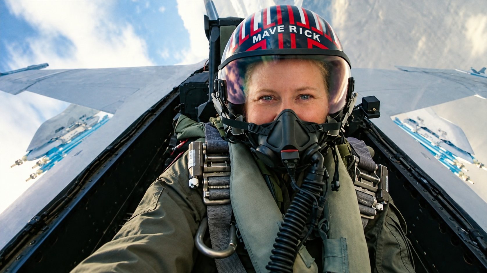 Erin in the cockpit of a fighter jet, wearing a flight helmet and oxygen mask, with the wings and blue sky behind her.